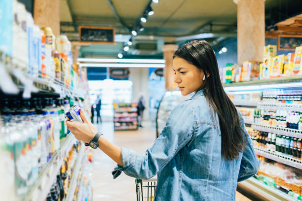Young woman buying beer or ale alcohol in liquor store, standing in front of shelf, holding can reading label